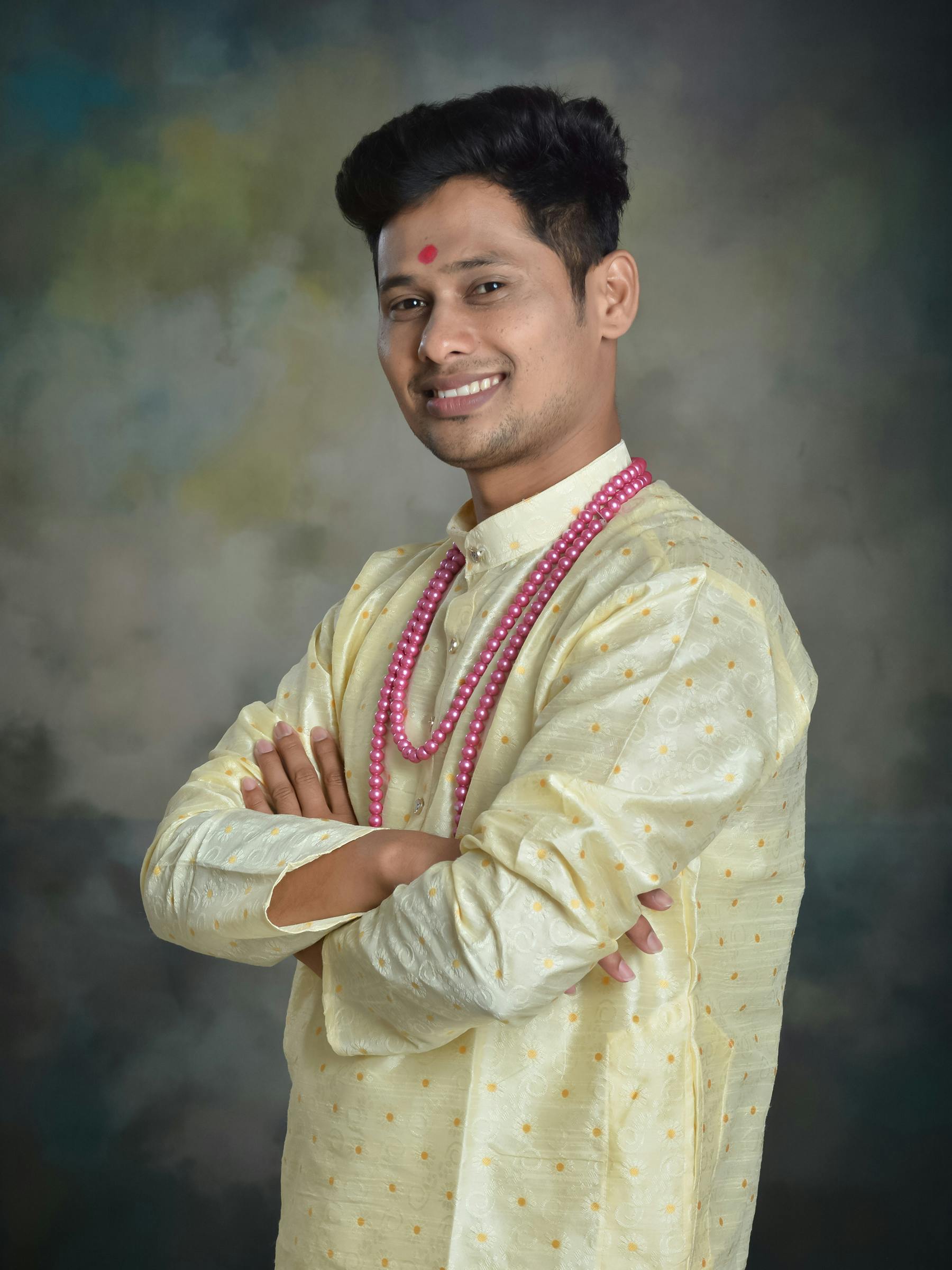 Smiling man in traditional Indian attire with a neutral background, arms crossed.