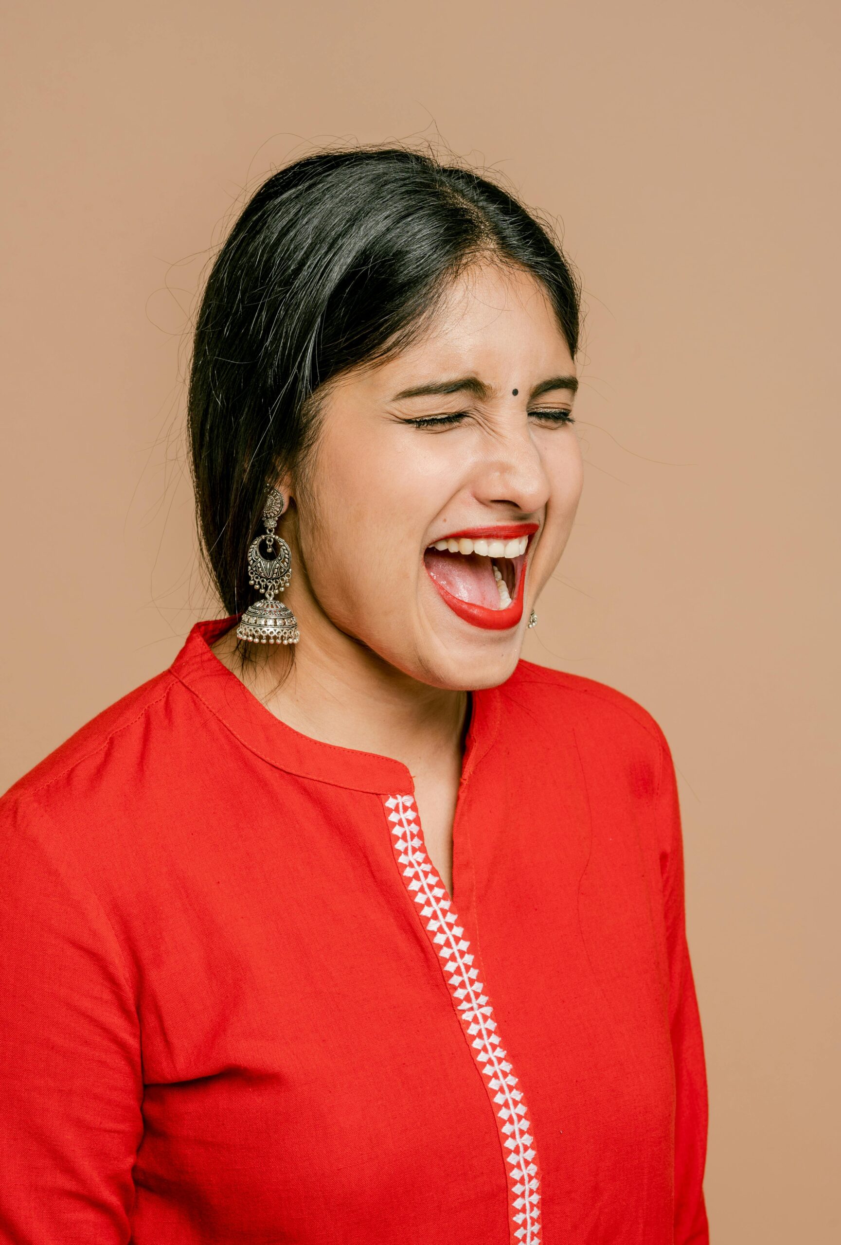 Close-up of a joyful Indian woman in a red dress expressing excitement with eyes closed and mouth open against a beige background.